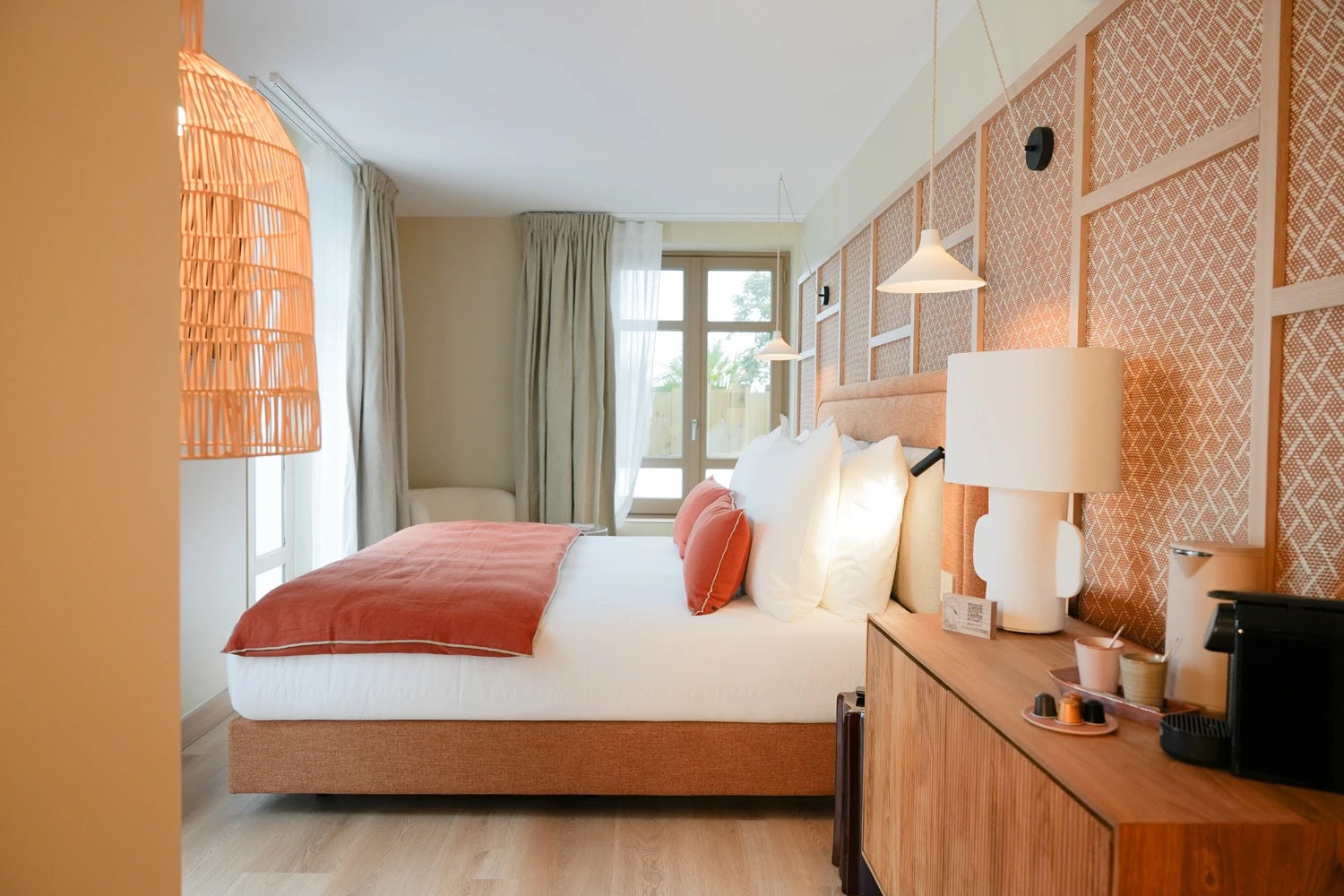 Chambre lumineuse avec un grand lit double habillé de linge blanc et un plaid orange, deux coussins orange, un mur décoré de panneau en osier, une commode en bois équipée d'une lampe blanche, d'une machine à café avec capsules, et des suspensions lumineuses en rotin près des fenêtres avec rideaux beiges.