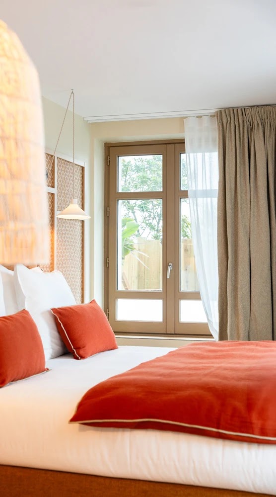 Chambre avec lit double décoré de coussins et couette orange, éclairage suspendu à gauche, fenêtre en bois avec rideaux beiges et voilage blanc laissant passer la lumière.