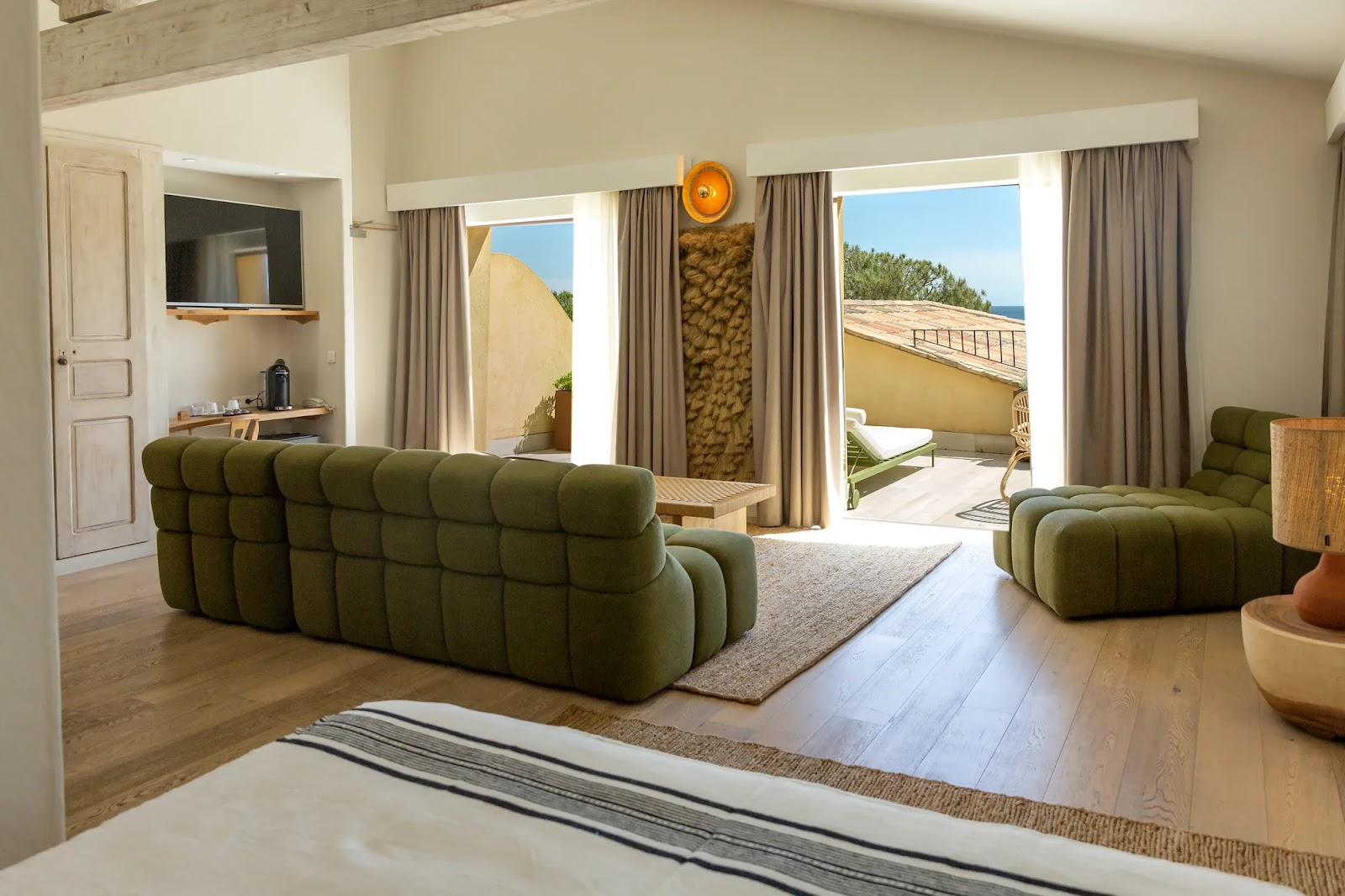 Chambre lumineuse avec parquet clair, canapé et fauteuil verts rembourrés, lit partiel en avant-plan, deux portes-fenêtres donnant sur une terrasse avec chaises longues et vue sur les toits et la mer.
