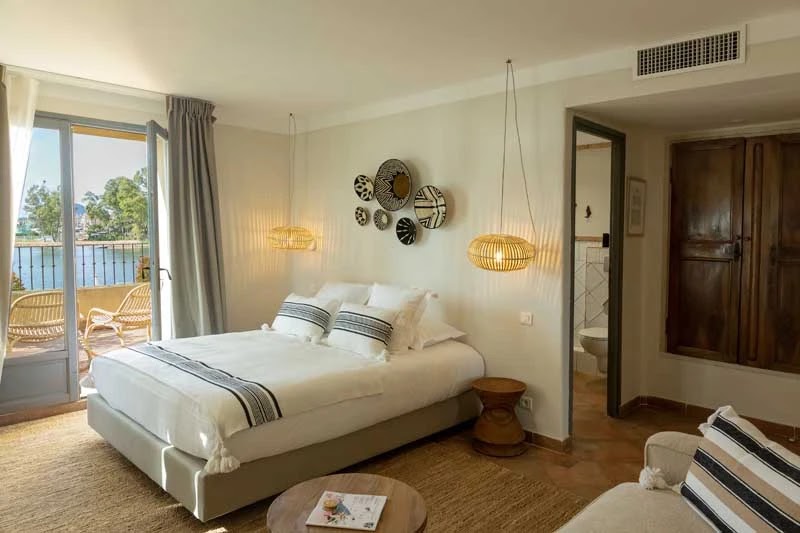 Chambre lumineuse avec grand lit double décoré de coussins blancs et noirs, deux luminaires suspendus en osier, petits meubles en bois, porte-fenêtre donnant sur balcon avec vue sur un plan d'eau et des arbres.
