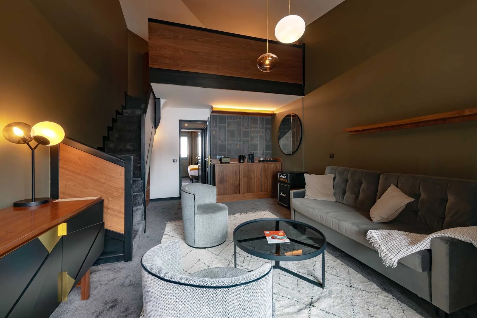 Salon moderne avec canapé gris foncé, deux fauteuils ronds, table basse ronde en verre, tapis clair à motifs, meuble bas avec lampe contemporaine à gauche, escalier avec marches couvertes de moquette, mur peint en vert olive, miroir rond au-dessus d'un meuble en bois avec appareils électroménagers, et mezzanine au-dessus.
