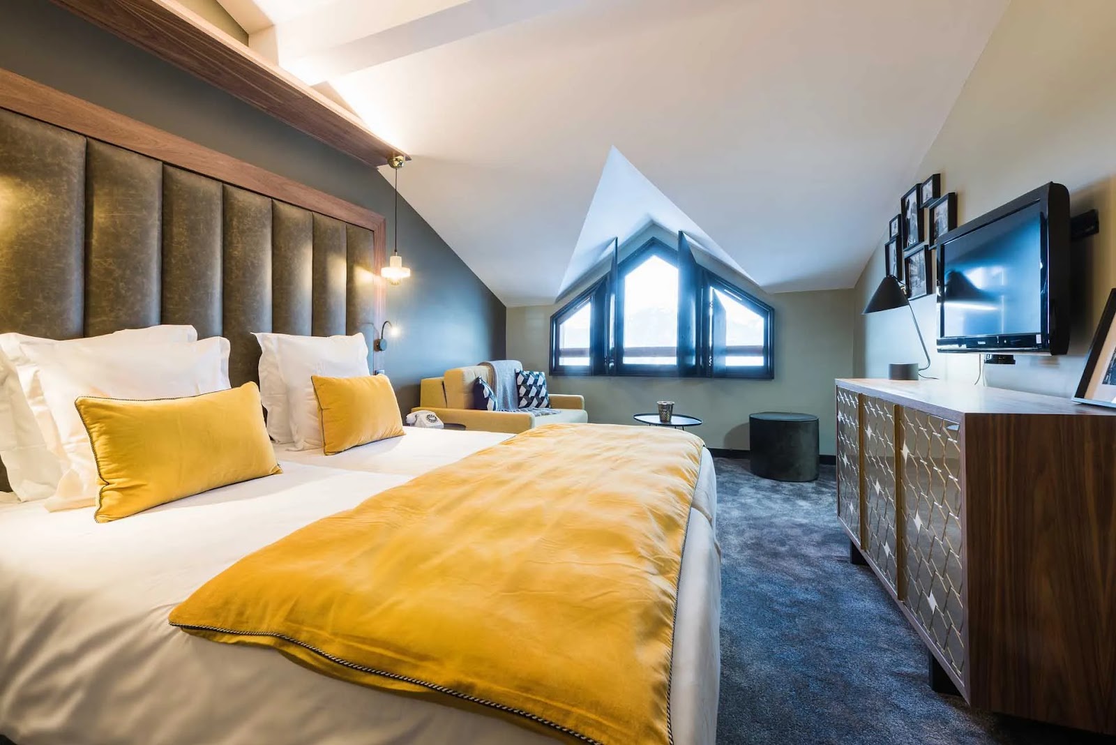 Chambre moderne avec grand lit double avec couverture et coussins jaunes, tête de lit en cuir, canapé beige près d'une fenêtre triangulaire, meuble bas en bois avec télévision murale et lampes.
