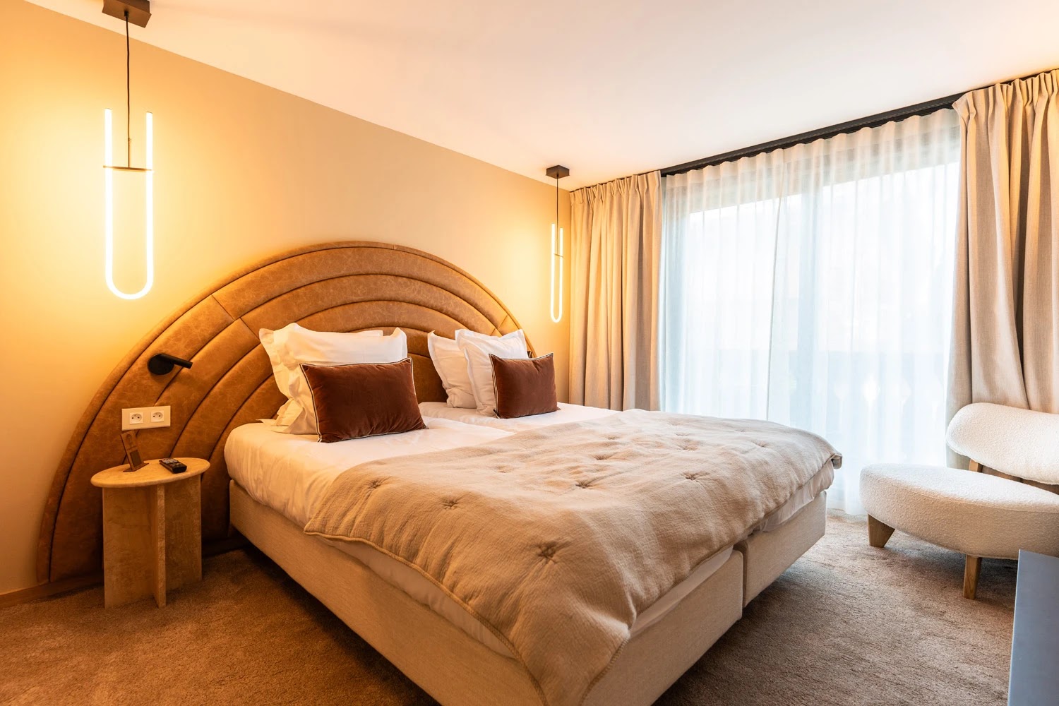 Chambre double moderne avec tête de lit en demi-cercle en cuir marron, lit avec couette beige et oreillers blancs et marron, suspensions lumineuses verticales de chaque côté, table de chevet en pierre, fauteuil beige à droite et grandes fenêtres avec rideaux beiges.