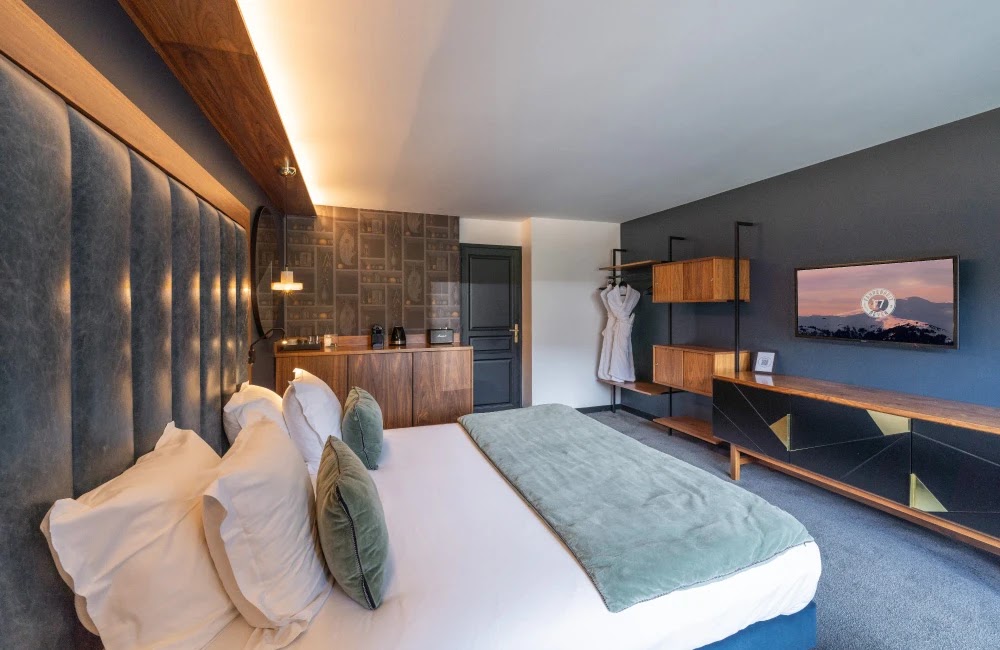 Chambre d'hôtel moderne avec grand lit double, tête de lit rembourrée, plusieurs oreillers blancs et verts, couverture verte, mur décoré avec papier peint à motifs, armoire en bois, peignoirs suspendus, meuble bas noir et bois avec télévision murale affichant une image de montagne au coucher du soleil.