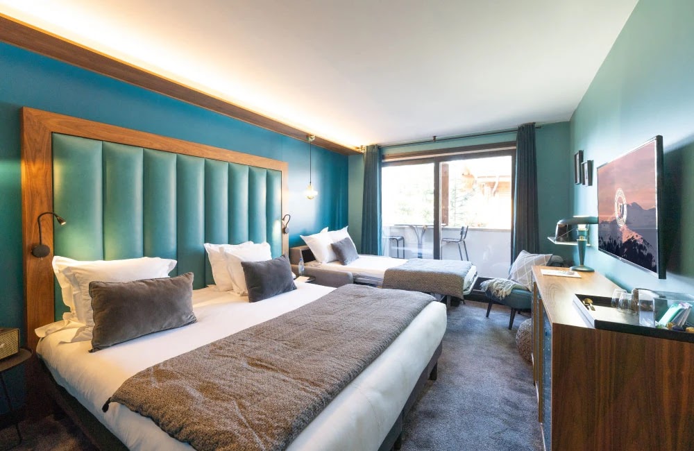 Chambre d'hôtel moderne avec un grand lit double et un lit simple, tête de lit bleue rembourrée, murs bleu-vert, télévision murale, et balcon avec vue extérieure.