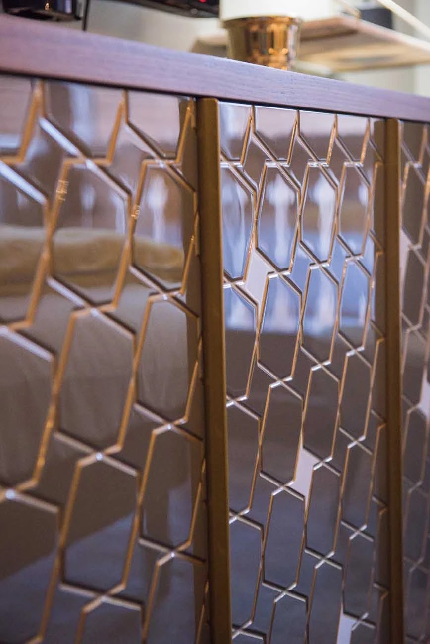 Gros plan sur un garde-corps décoratif avec un motif géométrique doré fait de formes hexagonales et rectangulaires, reflétant partiellement un canapé beige et un environnement intérieur flou.