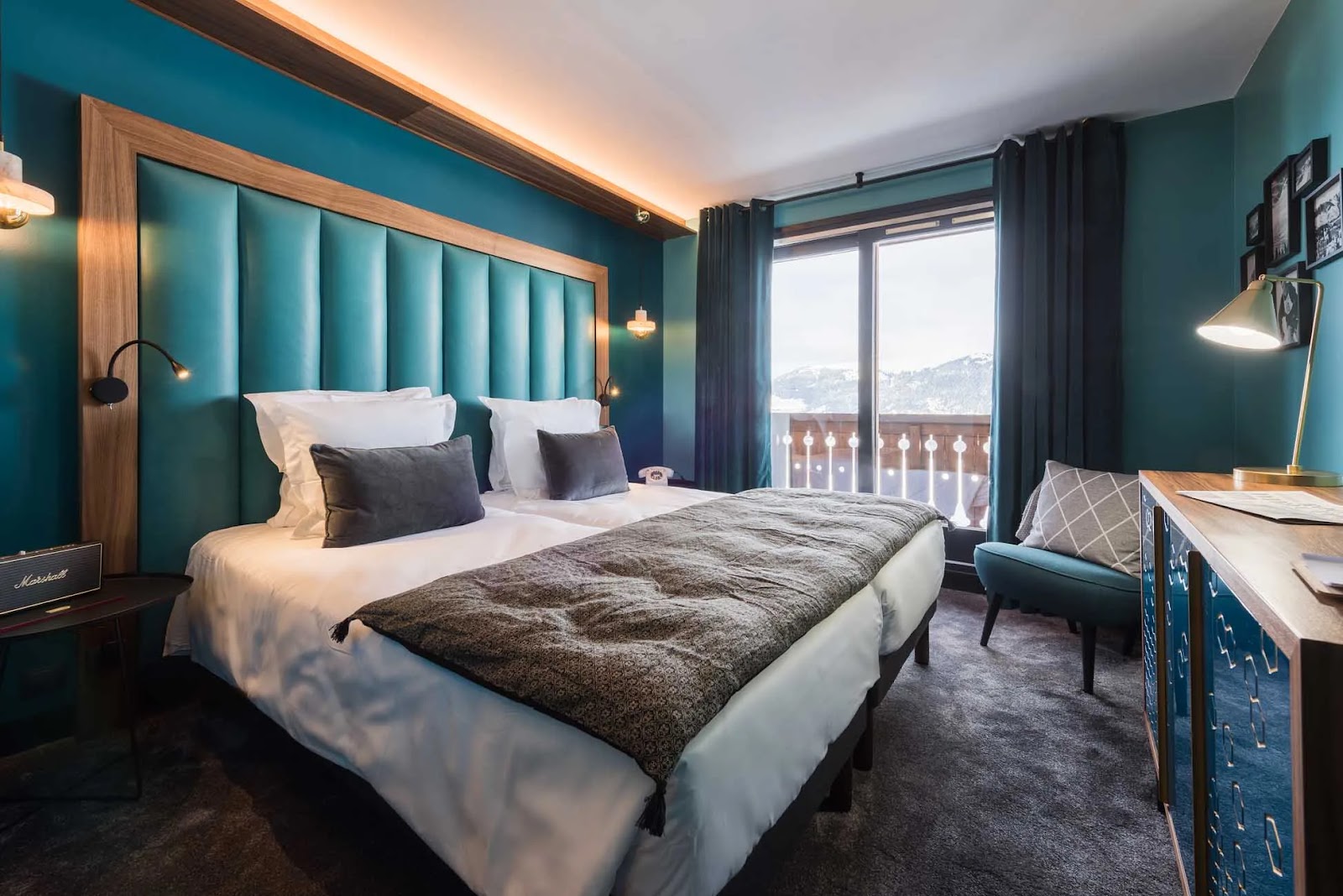 Chambre d'hôtel moderne avec un grand lit double bordé de coussins gris, une tête de lit rembourrée bleu canard encadrée de bois, un fauteuil bleu avec coussin, une armoire en bois et bleu, une lampe de bureau dorée, et une fenêtre donnant sur un balcon avec vue sur des montagnes enneigées.