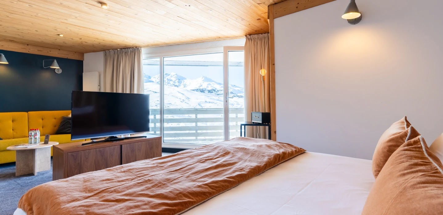 Chambre moderne avec un grand lit recouvert d'une couette marron et des coussins assortis, un téléviseur sur un meuble en bois, un canapé jaune et une grande fenêtre donnant sur un paysage de montagnes enneigées.