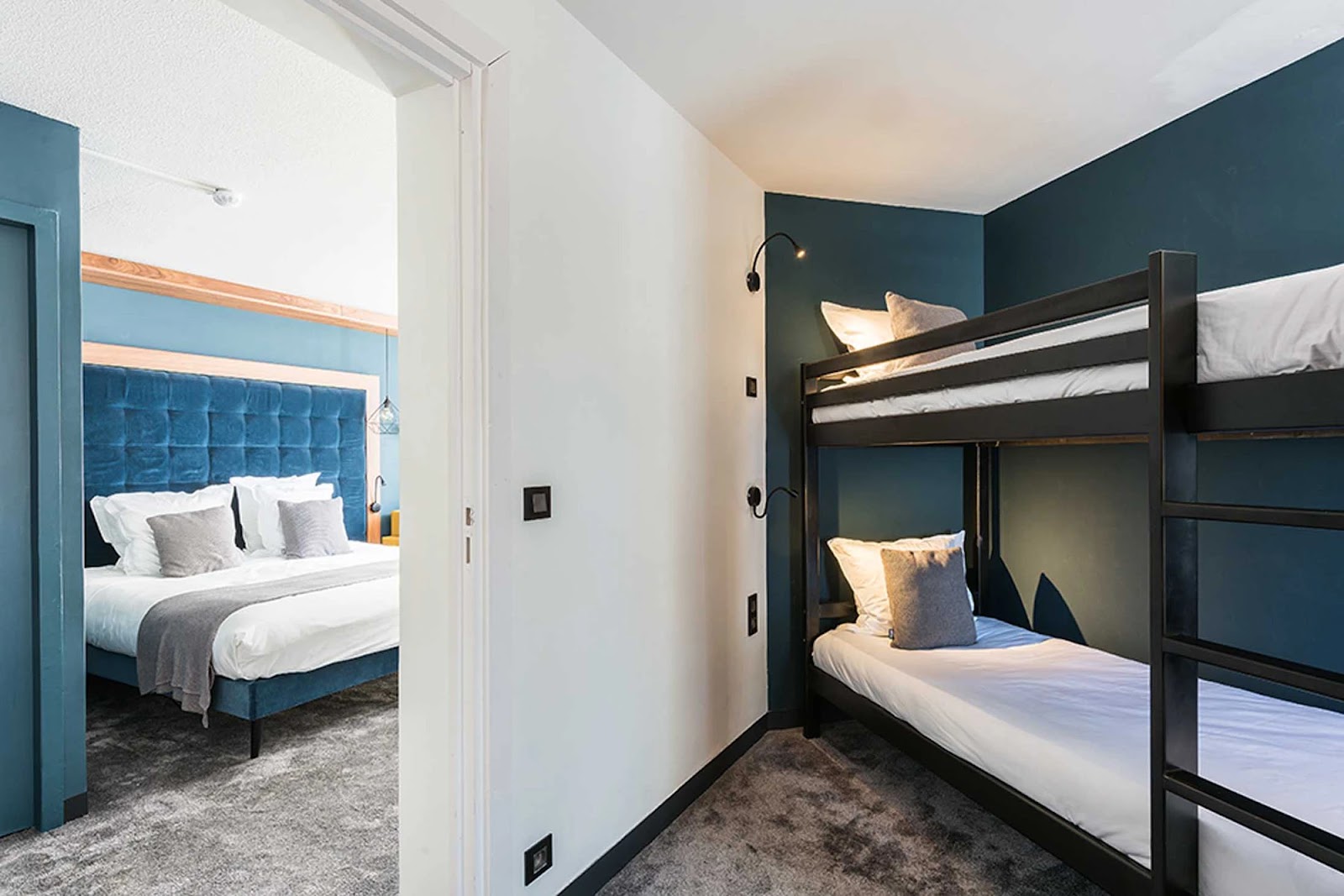 Chambre moderne avec un lit superposé noir contre un mur bleu foncé et un lit double avec une tête de lit bleu visible dans une pièce adjacente, le tout sur un sol moquetté gris.