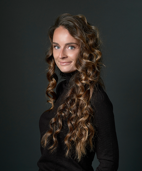 Portrait d'une femme souriante aux longs cheveux bouclés châtain clair portant un pull noir à col roulé devant un fond sombre.