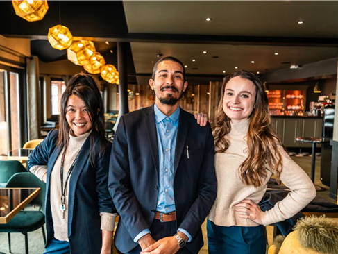 Trois personnes souriantes posent ensemble dans un intérieur de restaurant moderne avec des lampes suspendues dorées en arrière-plan, deux femmes encadrent un homme portant un costume bleu.