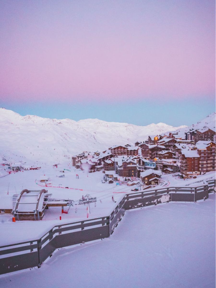 Village de montagne enneigé avec des chalets et immeubles sous un ciel au dégradé rose et bleu, avec une télécabine et des pistes de ski au premier plan.