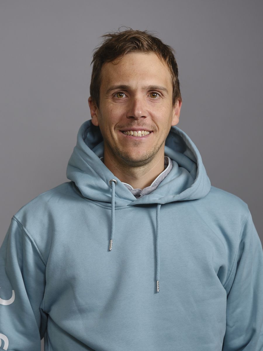 Homme souriant portant un sweat à capuche bleu clair contre un fond gris neutre.
