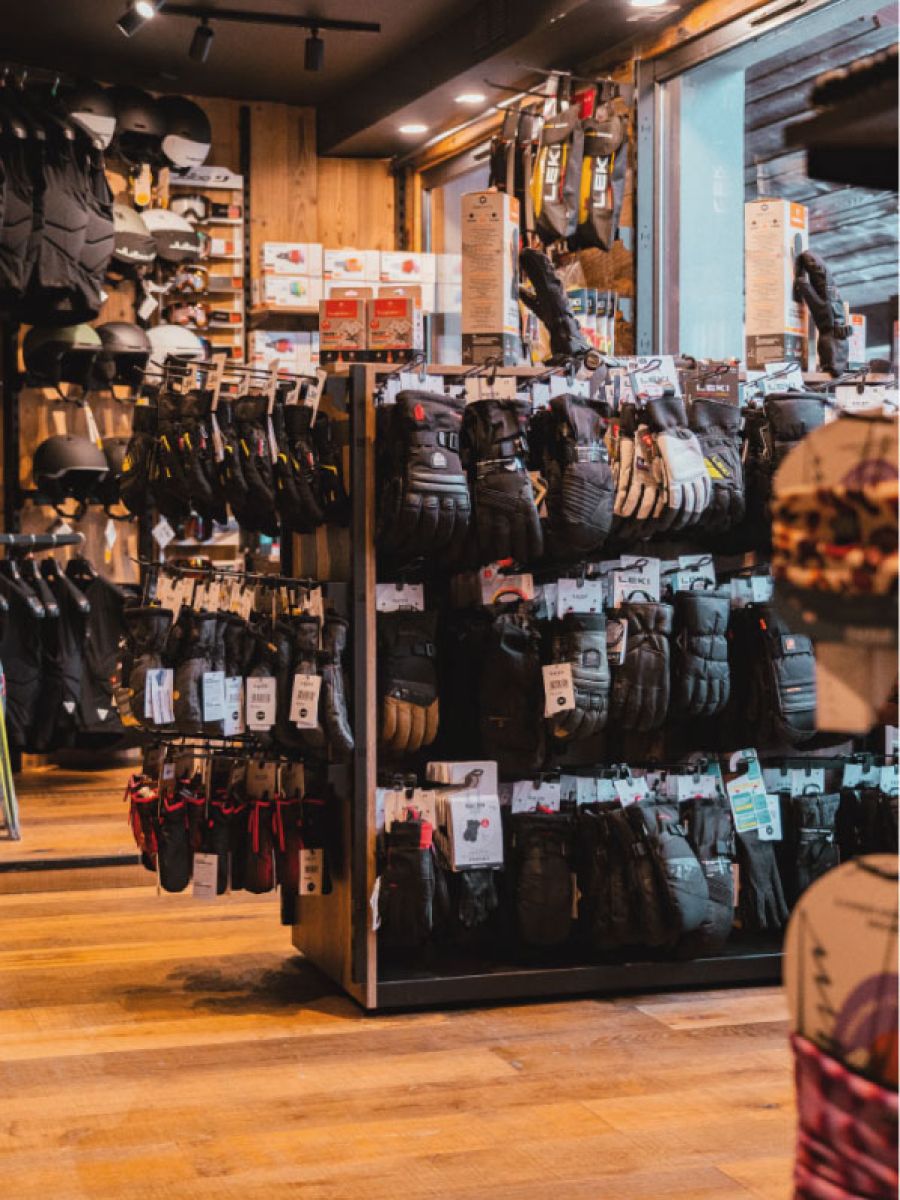 Intérieur d'un magasin de sport d'hiver avec des gants de ski exposés sur des présentoirs centraux et des casques accrochés au mur à gauche.