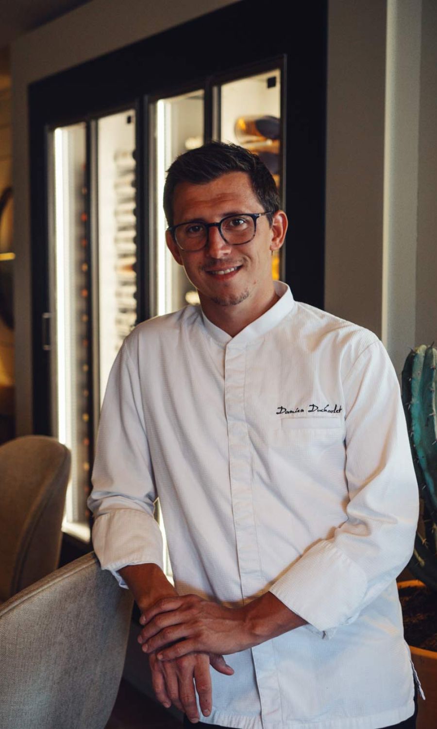 Homme portant une veste de chef blanche avec le nom Damien Ducharlet brodé, debout dans un intérieur avec un meuble vitré à bouteilles en arrière-plan, souriant et posant les mains l'une sur l'autre.