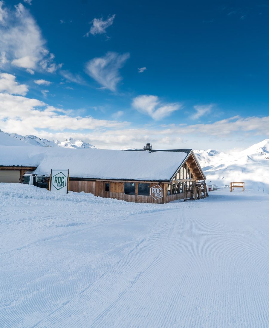 Bâtiment en bois nommé Roc Seven couvert de neige, situé dans un paysage montagneux enneigé sous un ciel bleu avec quelques nuages.