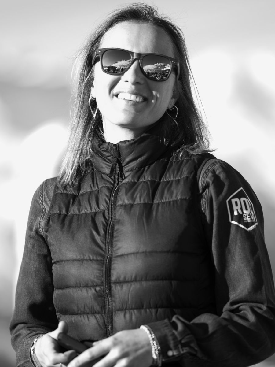 Portrait en noir et blanc d'une femme souriante portant des lunettes de soleil réfléchissant un paysage montagneux, un gilet matelassé et des boucles d'oreilles en forme de cœur.