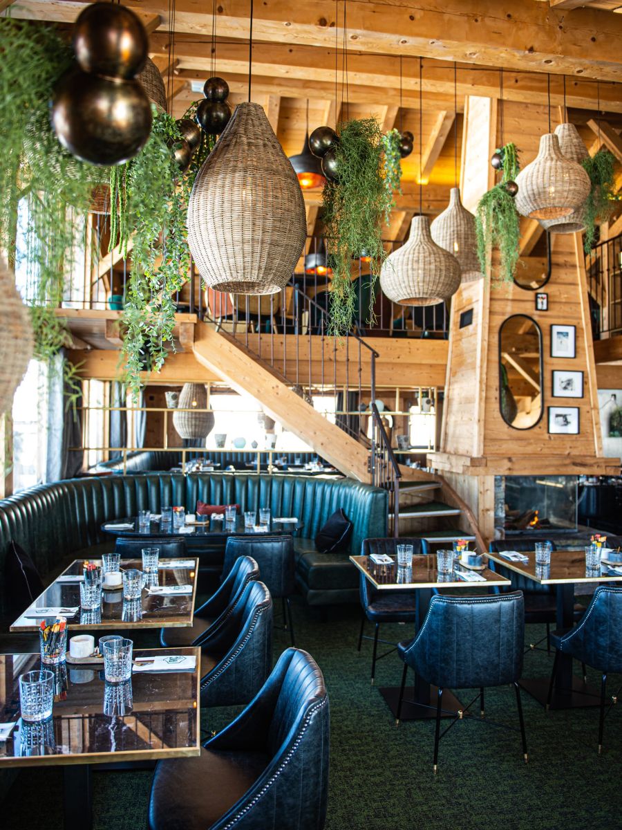 Intérieur d'un restaurant chaleureux en bois avec des suspensions en osier, des plantes vertes suspendues, des banquettes en cuir vert foncé et des tables dressées de verres et couverts, avec un escalier en bois menant à une mezzanine.
