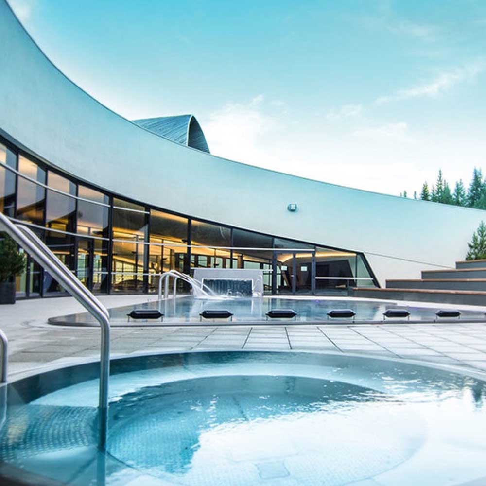 Vue extérieure d'un centre aquatique moderne avec deux bassins circulaires et rectangulaires, accompagnés d'escaliers en métal et d'une grande façade vitrée sous un ciel bleu clair.