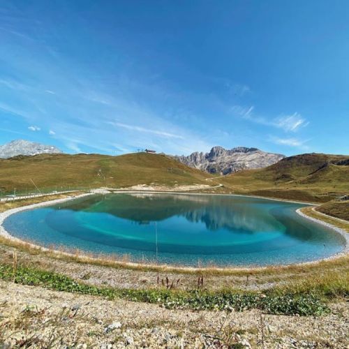 Petit lac circulaire aux eaux turquoises entouré de collines herbeuses avec des montagnes rocheuses en arrière-plan sous un ciel bleu clair.