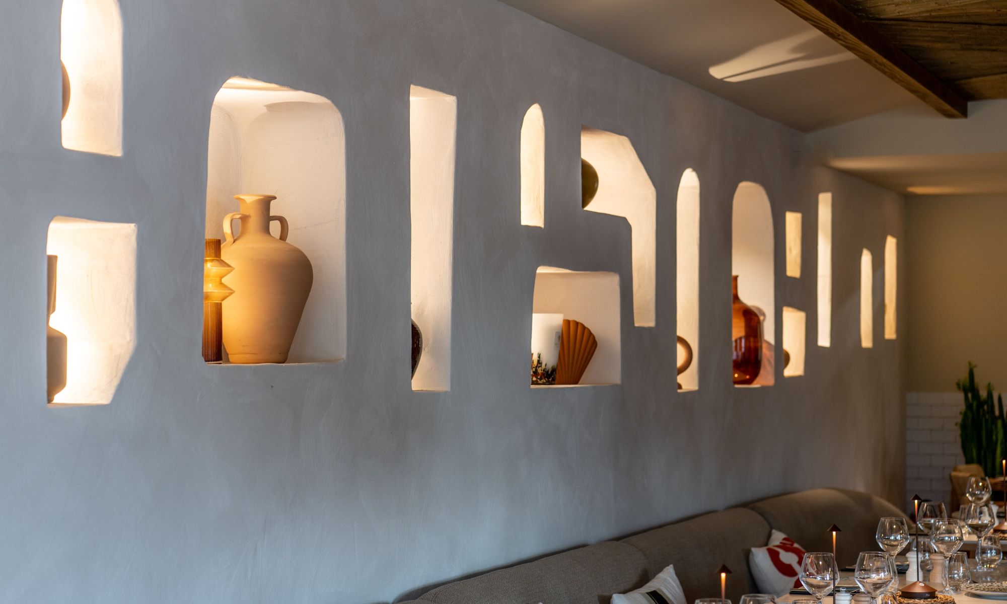 Mur intérieur avec niches éclairées contenant des vases décoratifs en céramique et du mobilier avec banquette grise et table dressée de verres et bougies.