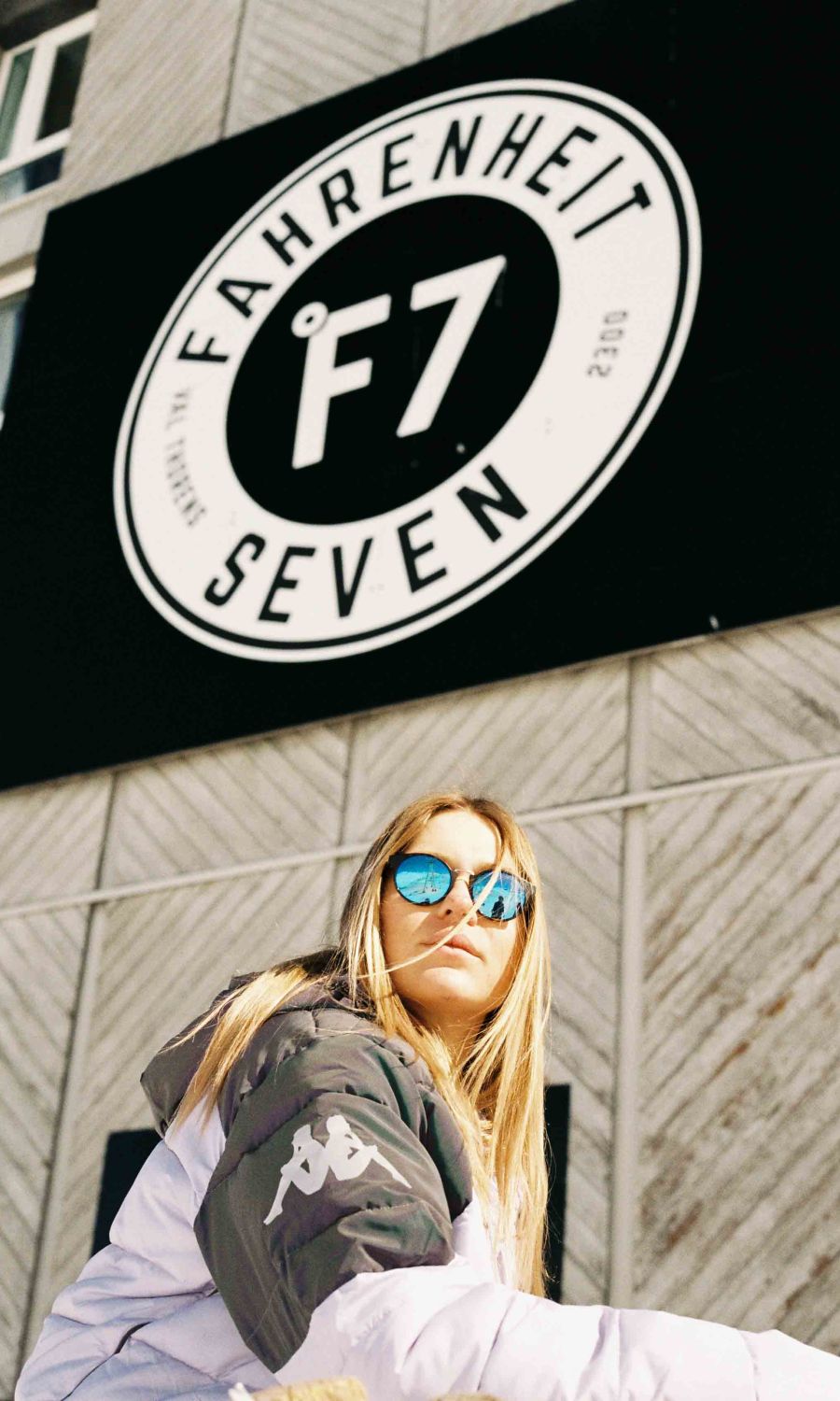 Une femme portant une veste blanche et noire et des lunettes de soleil réfléchissantes bleues pose devant un panneau circulaire noir et blanc avec le texte « Fahrenheit Seven F7 » sur un mur de bâtiment gris.