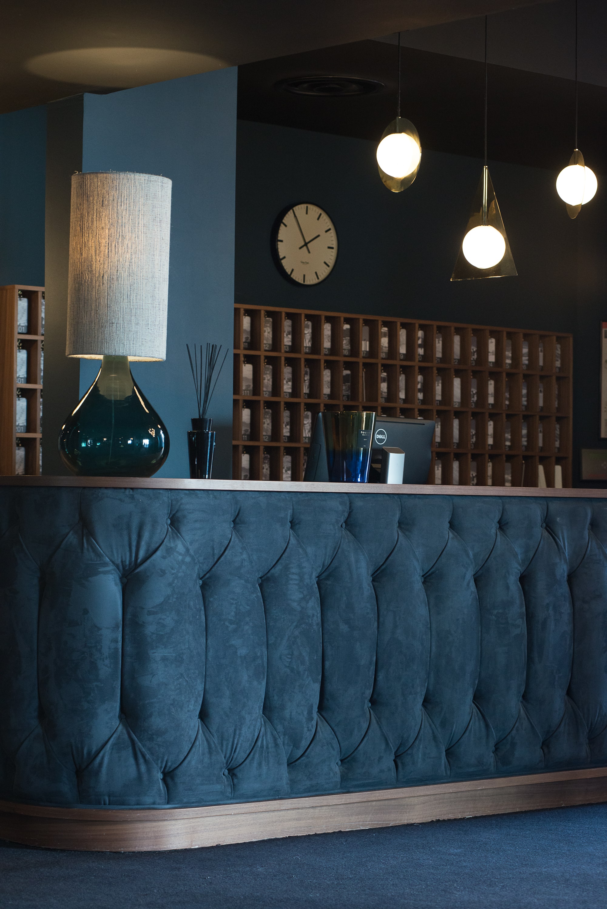 Accueil d'hôtel avec un comptoir en velours bleu foncé, une lampe à grand abat-jour, des suspensions lumineuses et une horloge murale au-dessus d'un casier à clés en bois.