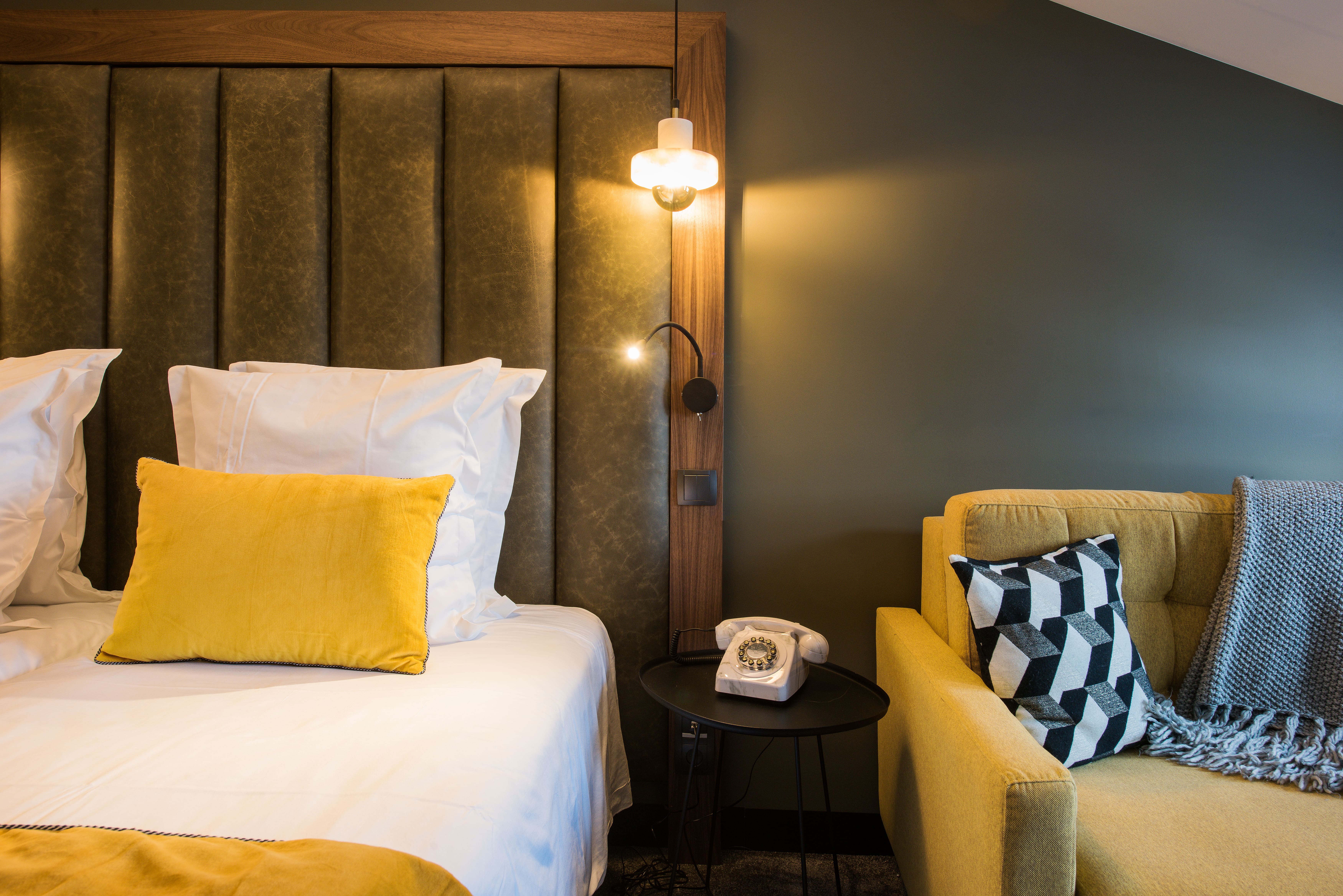 Chambre avec un lit blanc et un oreiller jaune, un mur vert, une table noire avec un téléphone rétro et un canapé jaune avec un coussin géométrique noir et blanc et une couverture grise.