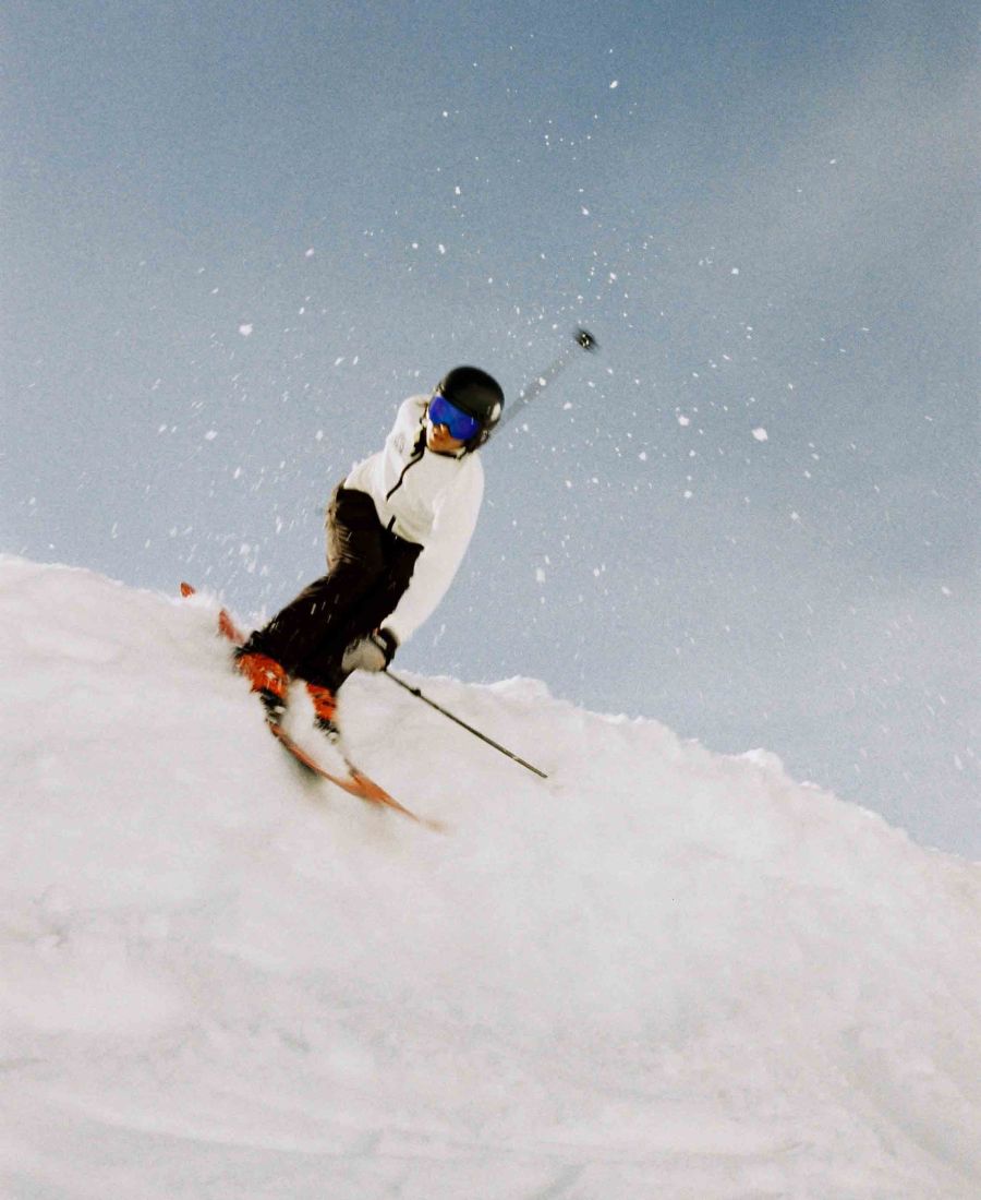 Personne qui descend une pente enneigée en skiant, portant un casque noir, des lunettes bleues et un vêtement blanc.