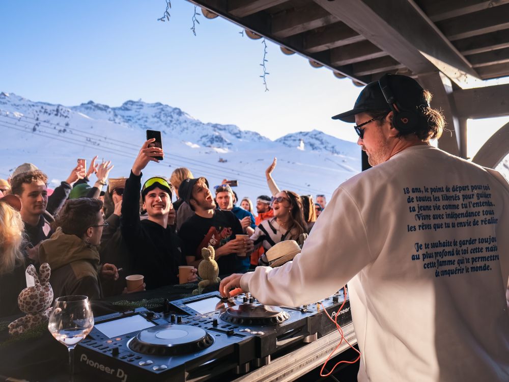 DJ portant un casque et des lunettes de soleil joue de la musique sur une table de mixage Pioneer devant un public enthousiaste à l'extérieur, avec des montagnes enneigées en arrière-plan.