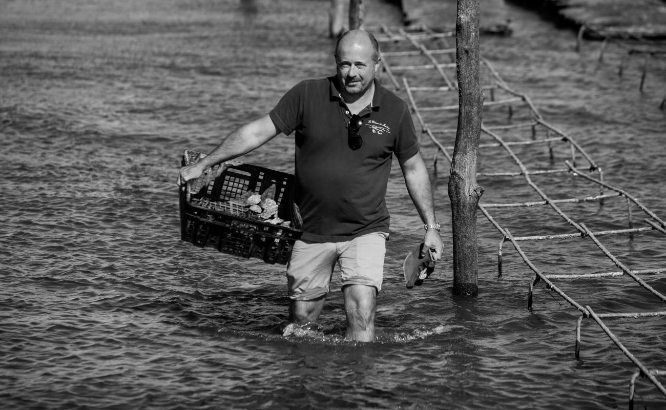 Un homme marche dans l'eau jusqu'aux genoux, portant un panier contenant des objets visibles et tenant des chaussures dans la main, avec une structure en bois partiellement immergée à côté de lui.