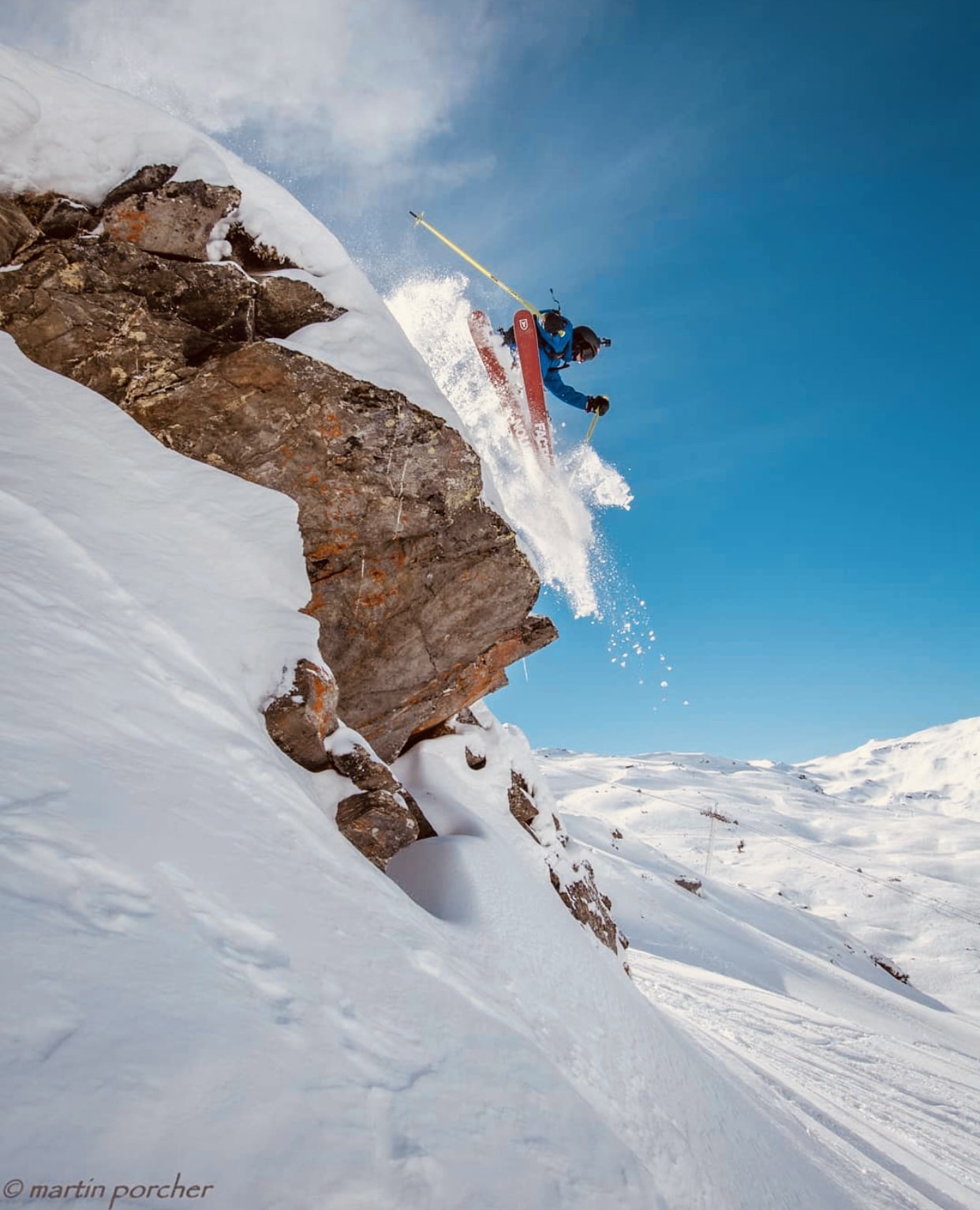 Skieur en tenue bleue sautant en hors-piste depuis une falaise rocheuse enneigée, projetant de la poudreuse sous un ciel bleu clair dans un paysage de montagne enneigée.