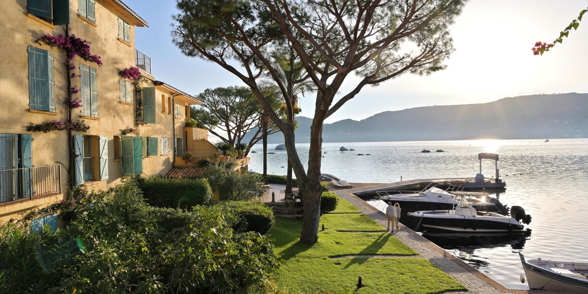 Vue d'un bâtiment jaune clair avec volets bleus entouré de plantes et arbres au bord d'un lac paisible où des bateaux blancs sont amarrés à un quai; deux personnes marchent le long du quai au coucher du soleil avec des montagnes en arrière-plan.