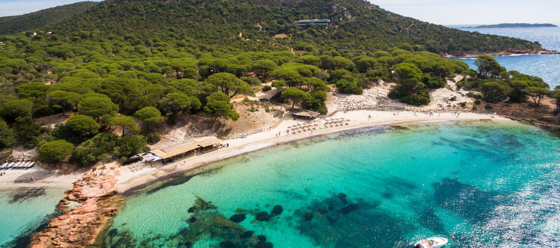 Vue aérienne d'une plage de sable fin bordée d'une mer turquoise claire avec quelques rochers, une petite barque blanche et un bois dense de pins sur une colline derrière la plage où se trouvent des parasols et un petit bâtiment en bois.