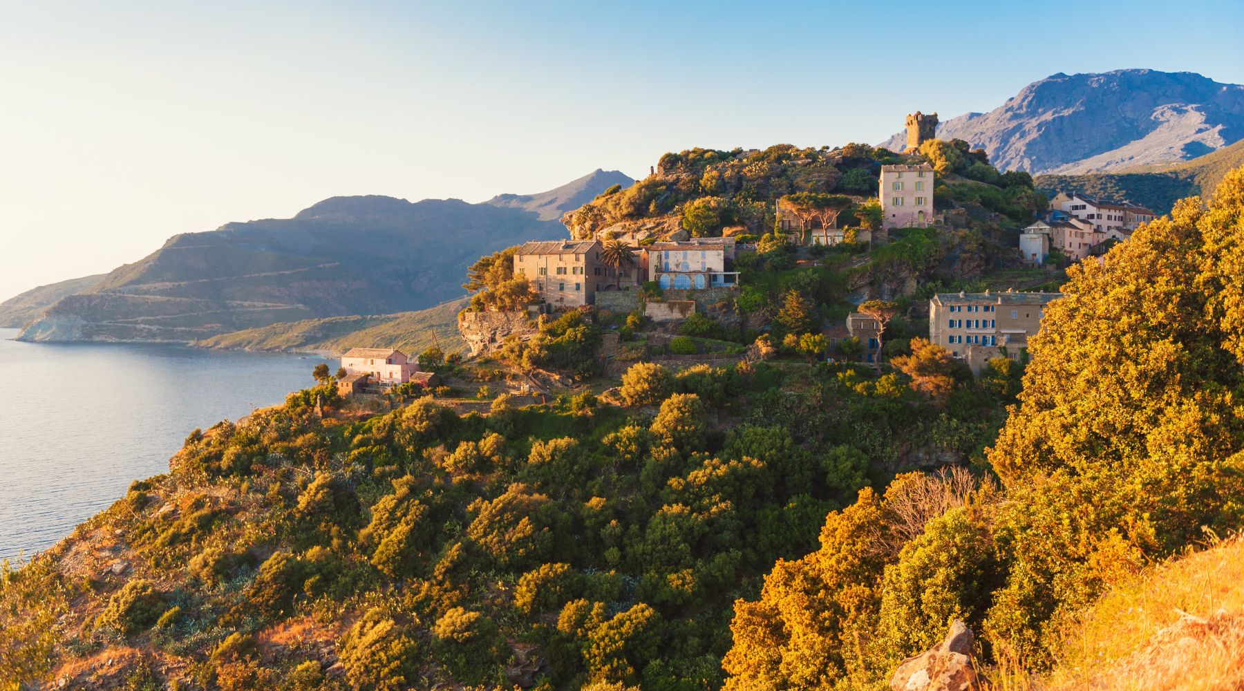 Village perché sur une colline verdoyante éclairée par le soleil couchant, avec des maisons en pierre et une tour ancienne, au bord d'une mer calme et entouré de montagnes en arrière-plan.