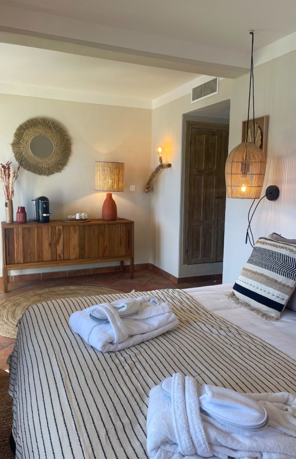 Chambre d'hôtel au style naturel avec un grand lit recouvert d'une couverture rayée beige et noire, deux peignoirs blancs pliés avec des chaussons posés dessus, coussin décoratif rayé beige, noir et blanc, lampe suspendue en osier à côté du lit, commode en bois avec une lampe à l'abat-jour en fibres naturelles, miroir rond décoratif en fibres, cafetière et objets de décoration sur la commode, murs clairs et porte en bois.