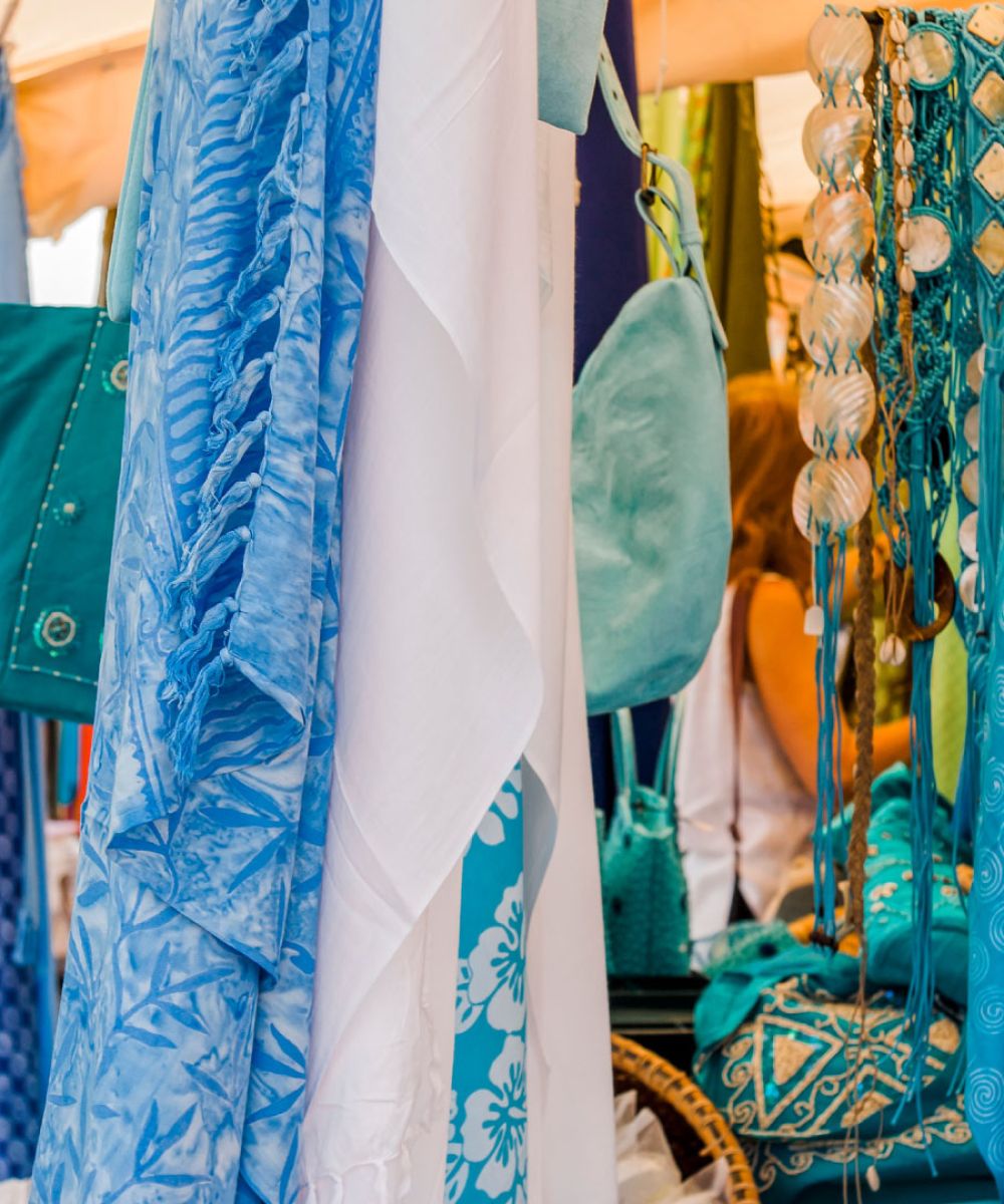 Écharpes et tissus bleus et blancs suspendus, avec sacs et accessoires turquoise exposés dans un marché, une personne floue en arrière-plan.