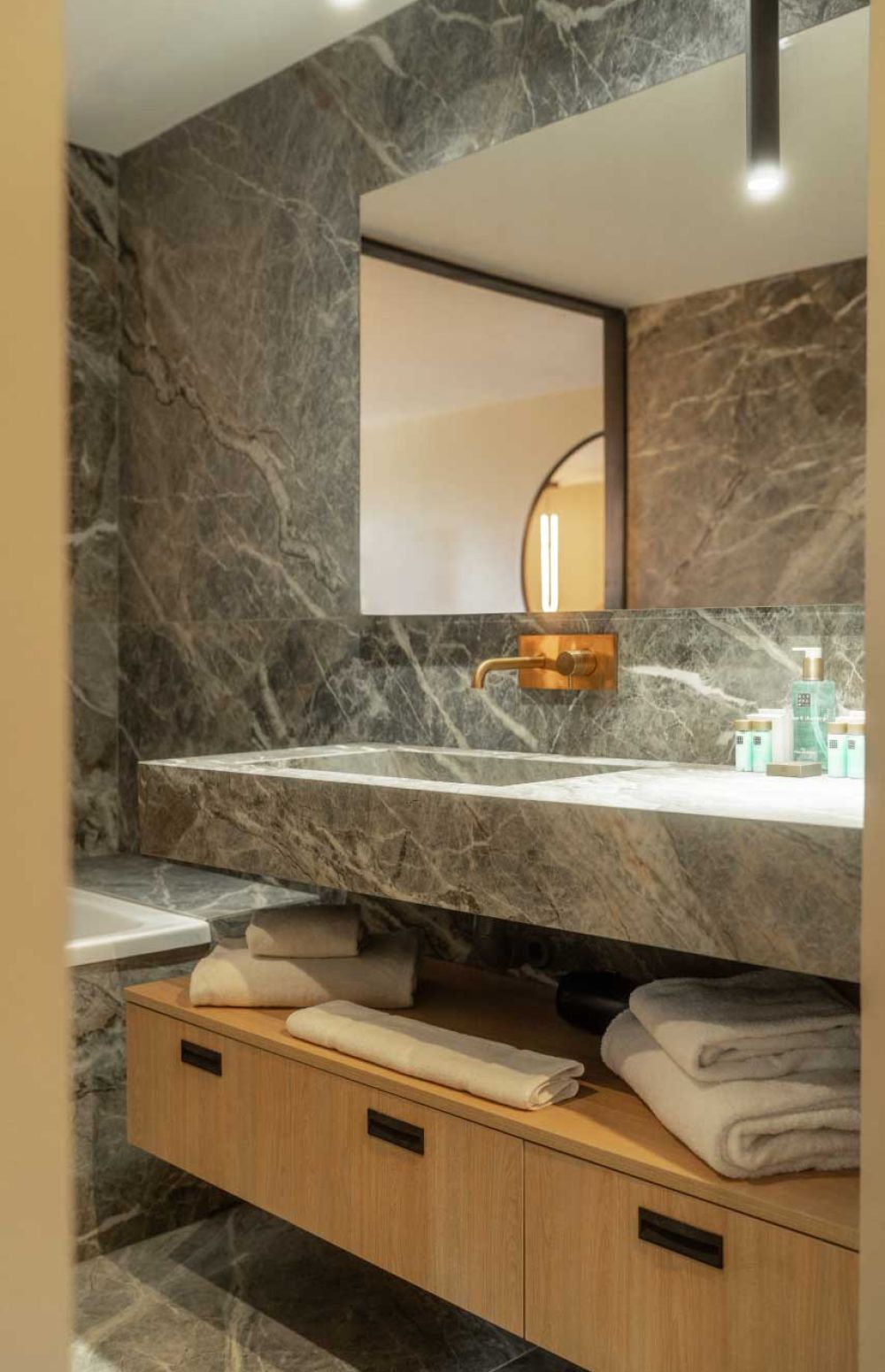 Salle de bain moderne avec lavabo et plan de travail en marbre gris, robinet doré mural, miroir rectangulaire, étagère en bois claire sous le lavabo avec serviettes blanches pliées et produits de toilette disposés sur le plan de travail.