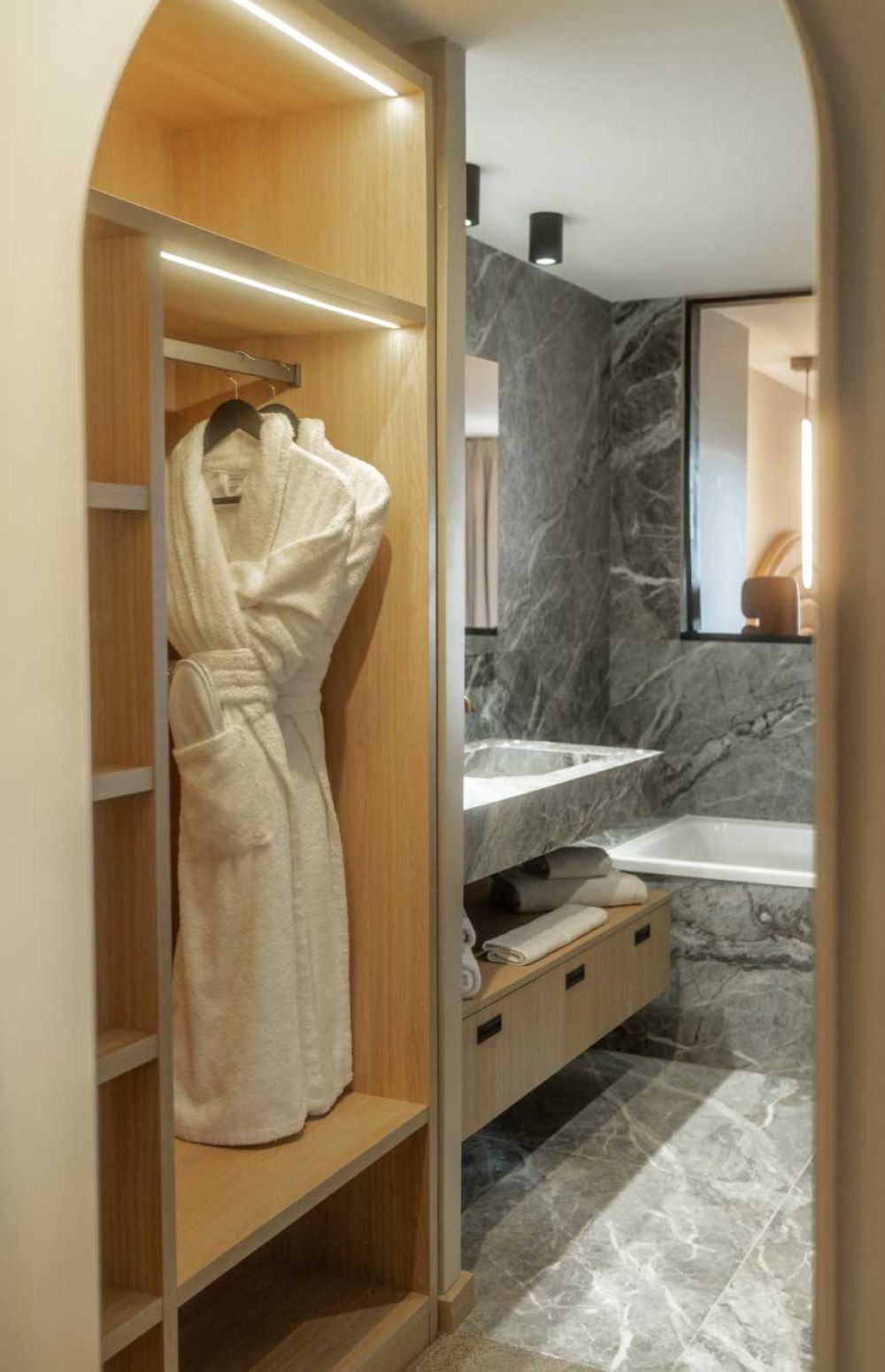 Vue d'une salle de bain d'hôtel moderne avec un peignoir blanc suspendu dans une armoire en bois éclairée à LED, un lavabo et une baignoire en marbre gris avec des serviettes pliées en dessous.