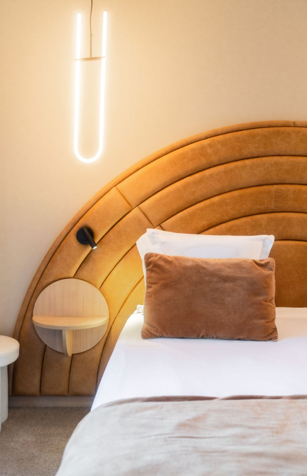 Chambre avec un grand lit à tête de lit ronde et capitonnée en cuir marron clair, un coussin rectangulaire marron, un coussin blanc, une lampe suspendue moderne blanche en forme de U et une petite tablette ronde en bois fixée au mur.