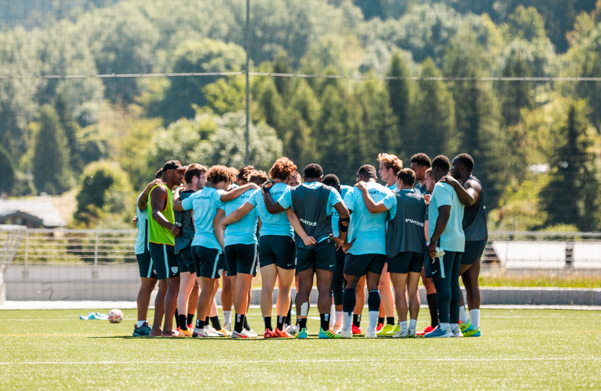 Une équipe de football en cercle sur un terrain herbeux lors d'une séance d'entraînement en plein air, avec des joueurs portant des maillots bleus et des shorts noirs, entourés d'arbres verts en arrière-plan.