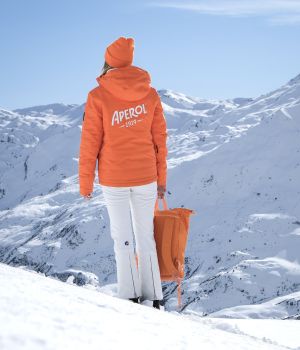Personne debout de dos sur une pente enneigée, vêtue d'une veste et d'un bonnet orange avec l'inscription Aperol, tenant un sac à dos orange, face à un paysage de montagnes enneigées sous un ciel bleu clair.