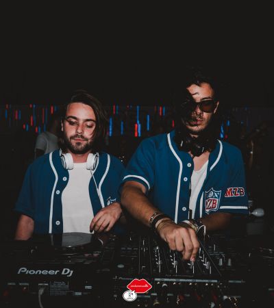 Deux DJ portant des maillots bleus jouent de la musique avec une table de mixage Pioneer DJ dans une ambiance sombre avec des lumières colorées en arrière-plan.
