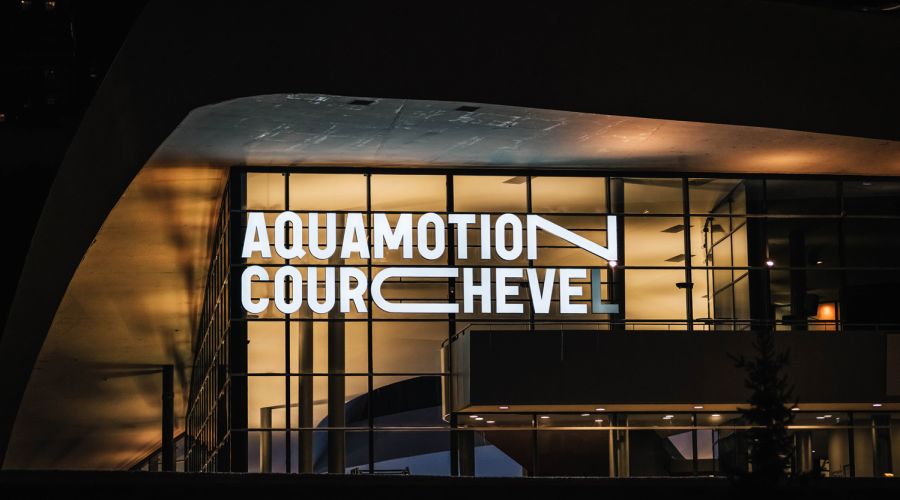 Façade nocturne éclairée d'un bâtiment moderne avec des grandes fenêtres vitrées et un panneau lumineux affichant le texte « AQUAMOTION COURCHEVEL ».