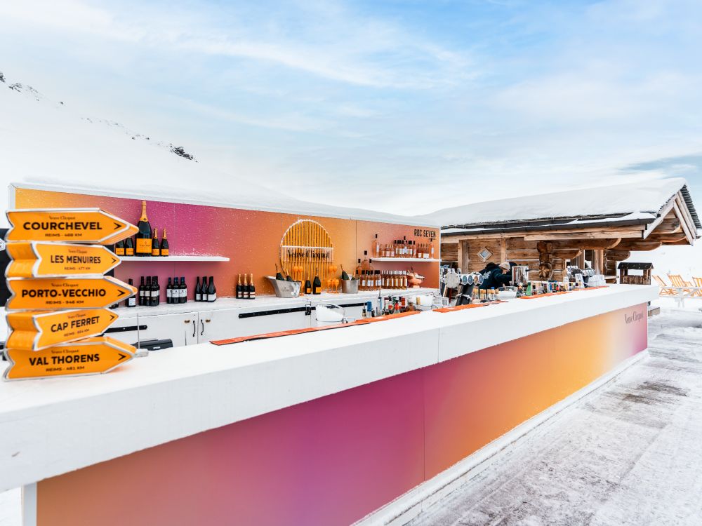 Bar extérieur avec comptoir long blanc et panneau dégradé rose et orange, bouteilles de champagne Veuve Clicquot exposées, enseigne directionnelle jaune indiquant Courchevel, Les Menuires, Porto-Vecchio, Cap Ferret, et Val Thorens, et chalet en bois en arrière-plan sous un ciel bleu avec quelques nuages.