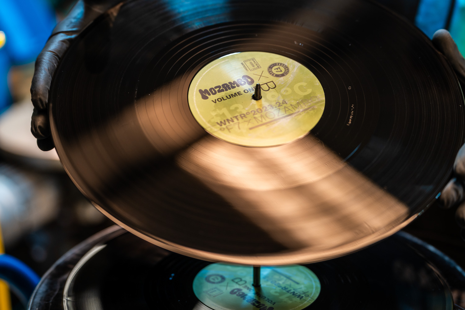 Main de personne portant un gant noir tenant un disque vinyle noir avec une étiquette centrale jaune portant les inscriptions « MOZAMBO VOLUME ONE », « WNTR°2023.24 » et un logo circulaire.