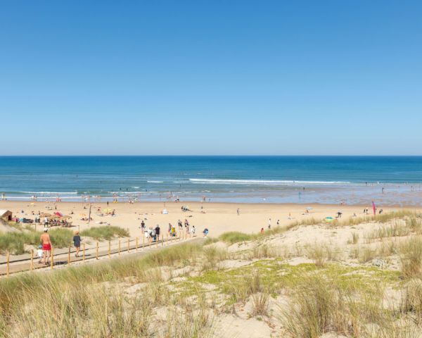 Plage sableuse avec des dunes herbeuses au premier plan, des gens se promenant et bronzant, et l'océan Atlantique calme sous un ciel bleu clair.