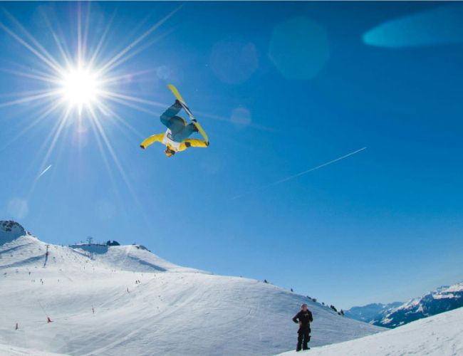 Skieur en combinaison jaune effectuant un saut acrobatique en l'air sous un ciel bleu clair avec un soleil éclatant dans une station de ski enneigée avec des montagnes en arrière-plan et une personne au sol observant.