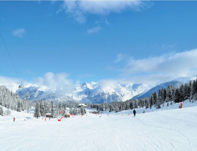 Paysage d'une station de ski avec neige abondante, skieurs sur une large piste, télécabines à gauche, sapins enneigés et montagnes enneigées en arrière-plan sous un ciel bleu avec quelques nuages.
