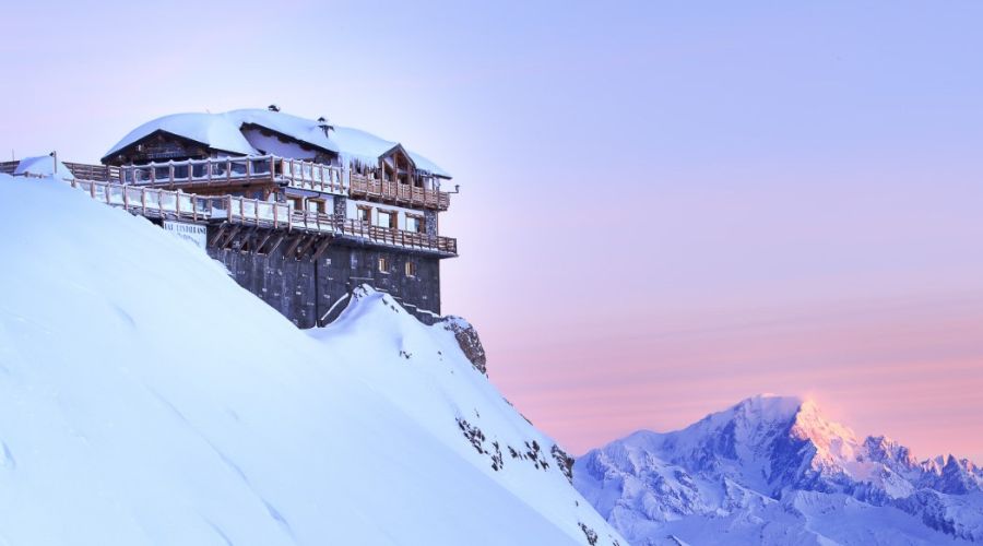 Chalet en bois perché au sommet d'une montagne enneigée au coucher du soleil, avec des montagnes enneigées en arrière-plan sous un ciel aux teintes roses et violettes.